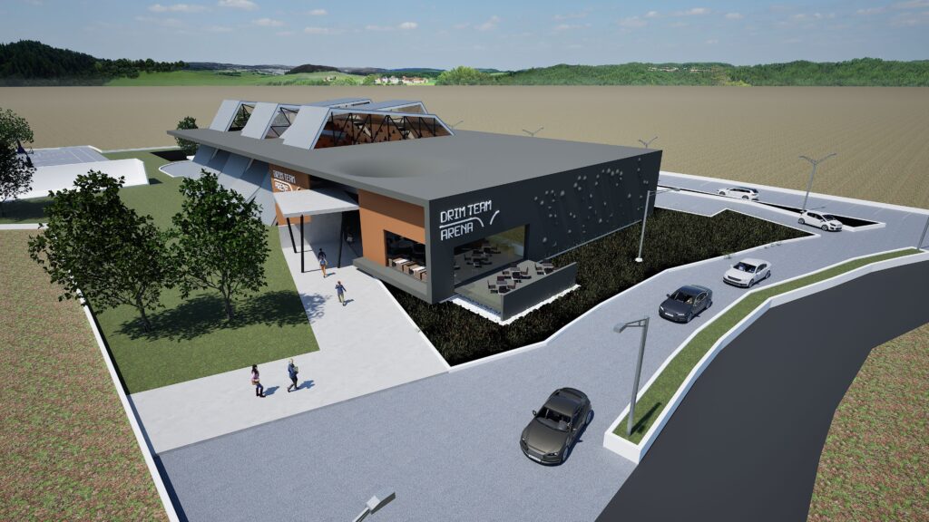 Aerial view of the Drimalas Basketball Academy, showcasing a contemporary sports facility with a sleek, low-profile design. The building features a combination of dark gray and white facades, accented by orange and a perforated panel section. Outdoor seating areas and landscaping surround the structure, with a parking lot and surrounding green fields visible in the background. The design emphasizes a modern aesthetic and integration with the natural landscape. By Modify Architects