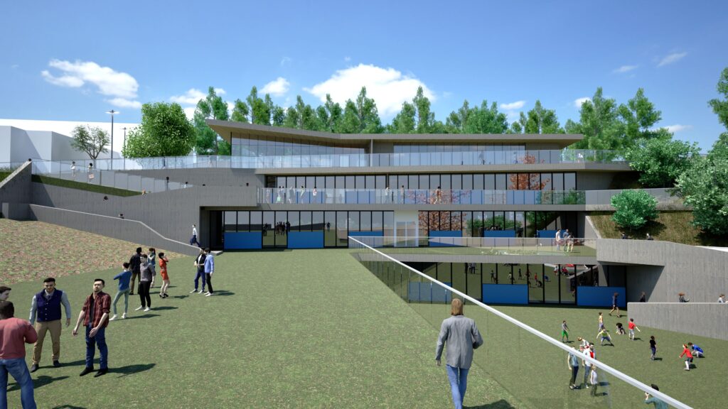 Exterior view of the Korifidis School, a modern two-story educational building integrated into a sloping green landscape. The building features a long, horizontal design with a glass facade and a curved roofline. Students and people are visible on the lawn and around the building, suggesting an active and welcoming environment. A fence runs in the foreground, and trees are visible in the background under a clear blue sky. By Modify Architects