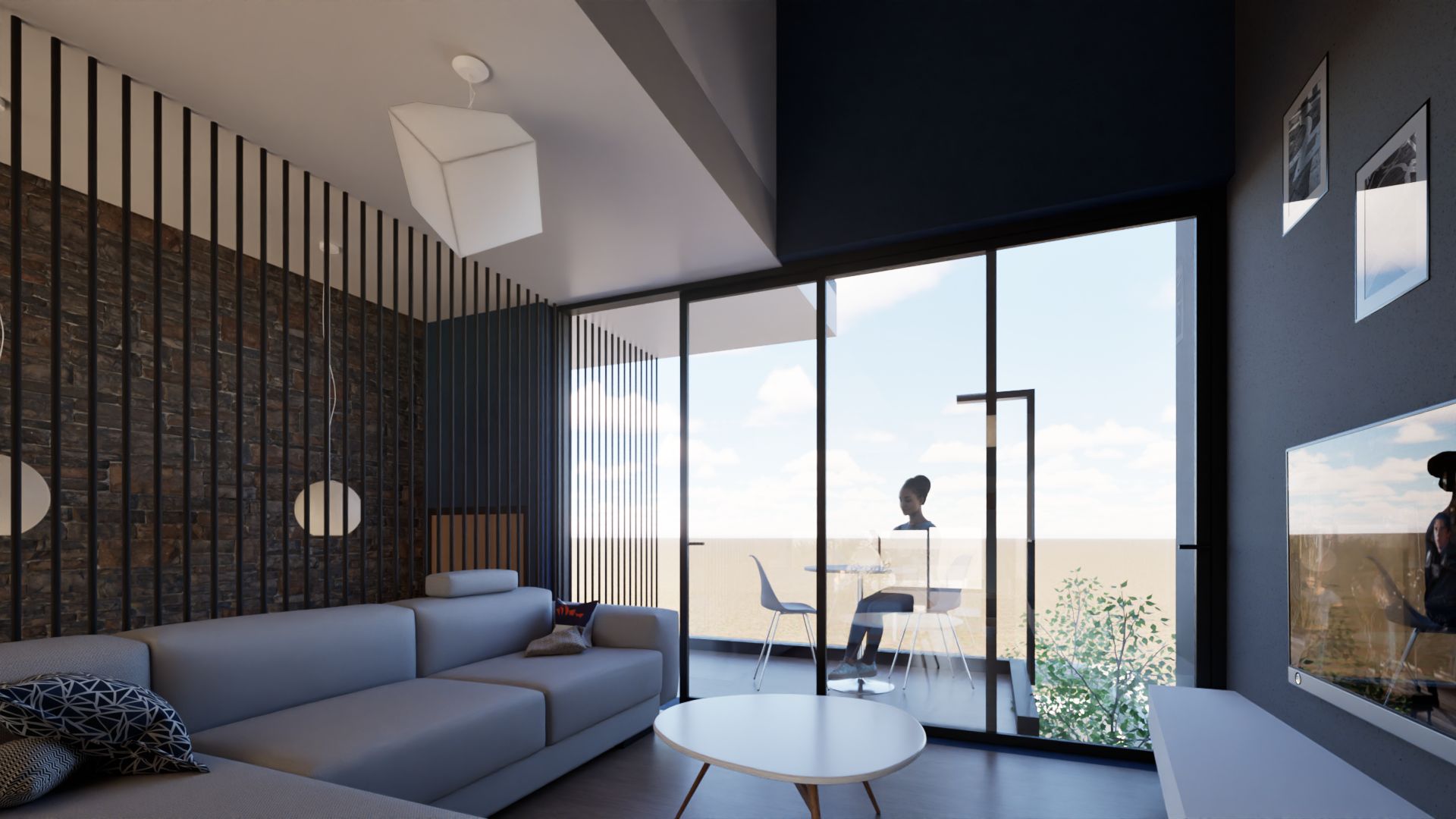 A bright, modern living room interior with a gray sectional sofa, a white coffee table, and large glass sliding doors leading to a balcony with a dining set and a scenic view of the sky and distant landscape. A brick accent wall and sleek lighting fixtures add to the contemporary design. By Modify Architects