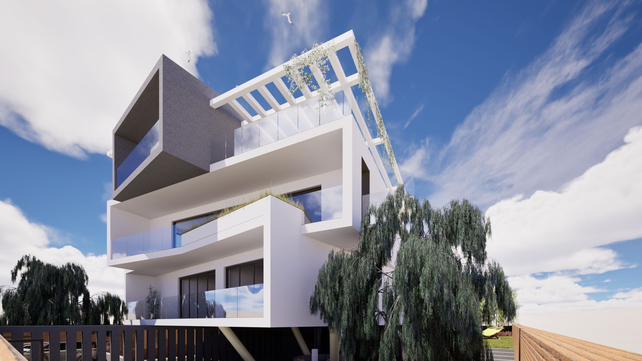 A low-angle exterior view of Residence 24, a modern white villa with a striking geometric design. The building features multiple levels, each with balconies enclosed by glass railings. A grey, angular section contrasts with the predominantly white facade, and greenery cascades from the upper levels, softening the architectural lines. The villa is set against a vibrant blue sky with fluffy white clouds, and surrounded by trees and landscaping. By Modify Architects